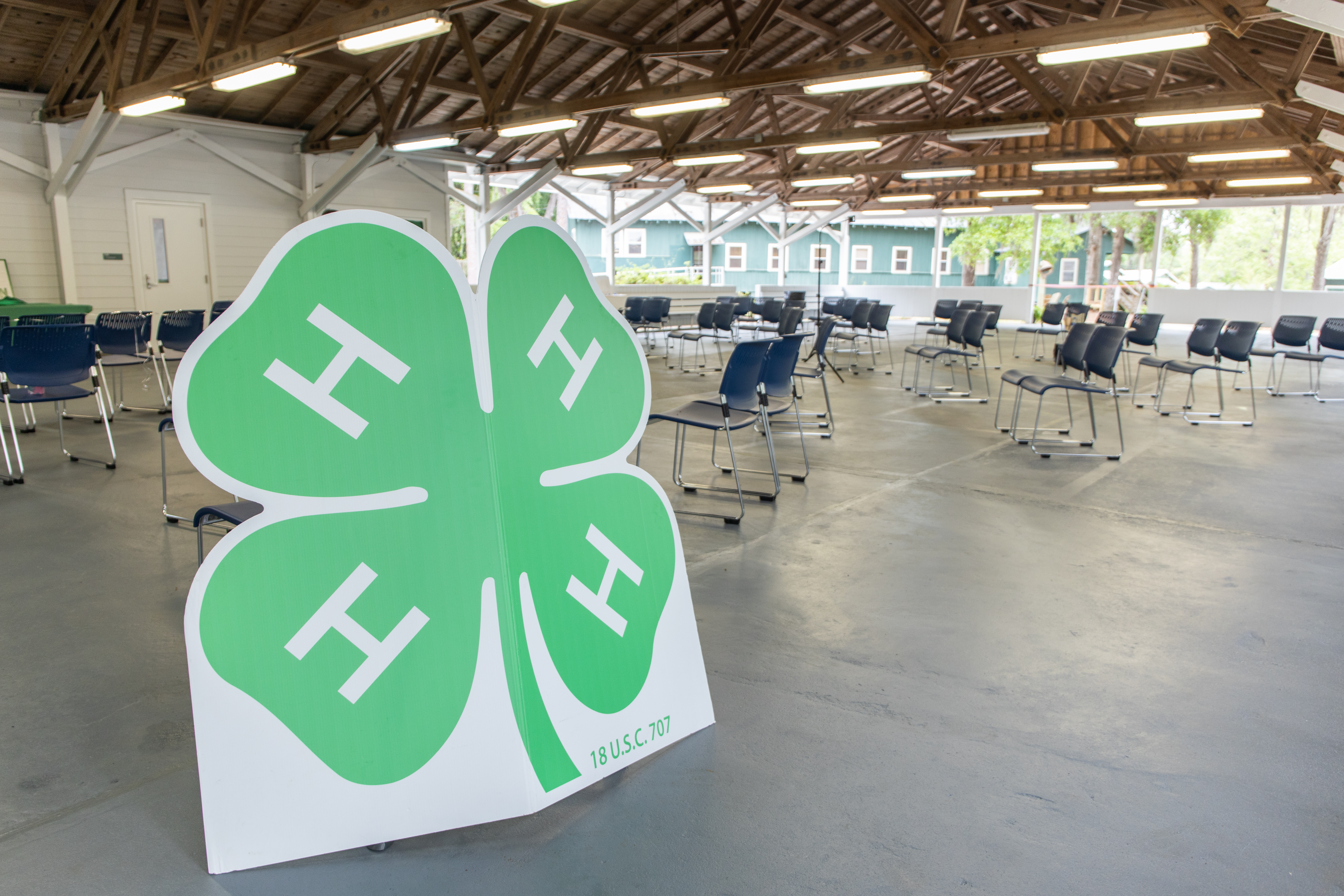 Logo of 4-H displayed prominently in a large, open pavilion with several circular tables and chairs arranged throughout the space. The structure has a wooden truss ceiling and is well-lit.