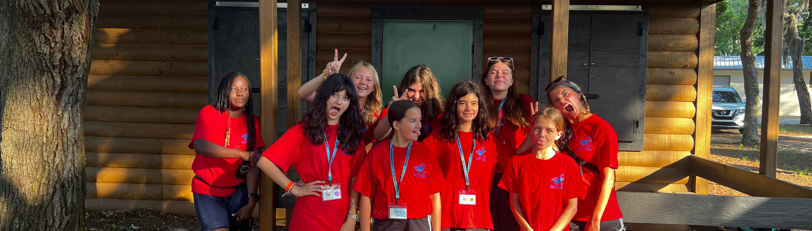 Banner image of youth smiling and posing in front of a camp cabin