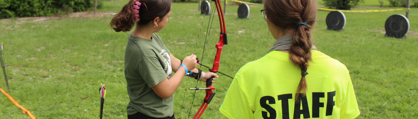 Photo of staff member at camp cloverleaf showing a youth how to properly hold a bow for archery