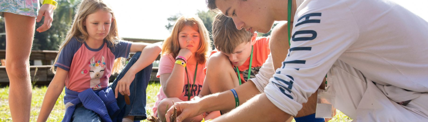 Web banner image of Group of children engaged in an outdoor educational activity, focusing intently as an instructor demonstrates a task.