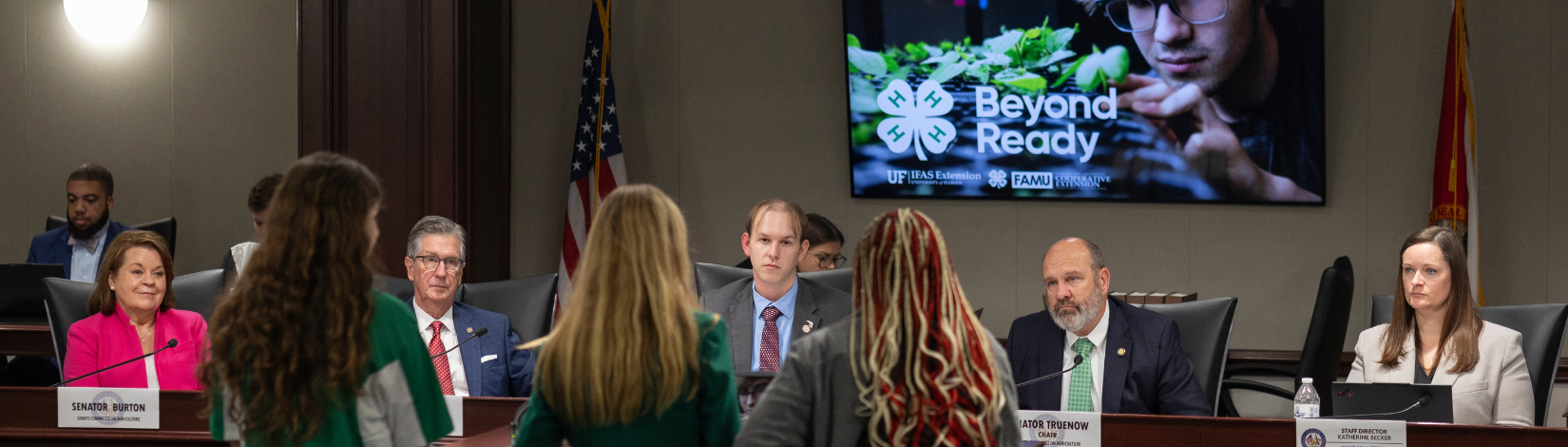 Meeting in progress at a conference table with nine participants, in front of a screen displaying 'Beyond Ready' by Milligan University, and a U.S. flag to the right.